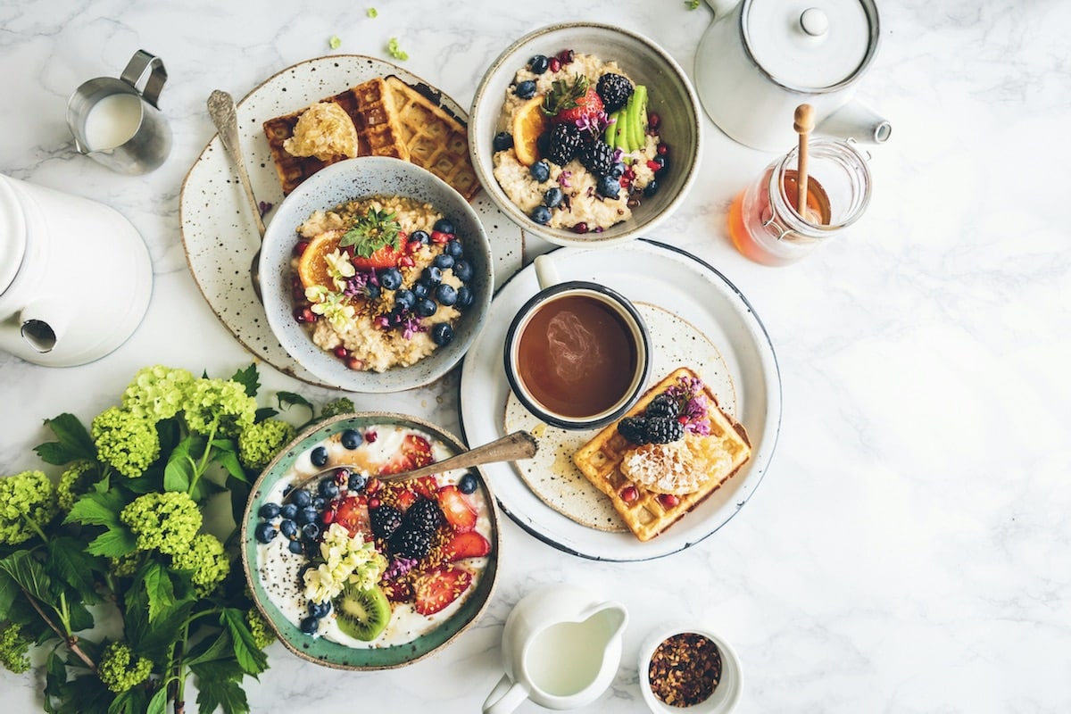 Breakfast spread, three bowls with oatmeal and various berries. Two plates with waffles, surrounded by decorative cups that contain milk, honey, and spices.